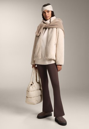 Kobieta w beżowej, teksturowanej kurtce na kremowym swetrze, brązowych rozszerzanych spodniach, z opaską na głowie, trzyma kremową pikowaną torbę, z beżowym swetrem zarzuconym na ramiona.