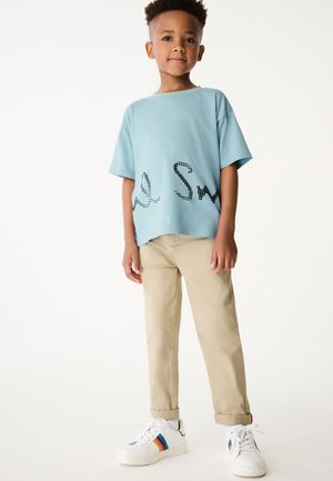 Gai zils īsām piedurknēm T-krekls ar melniem punktveida burtiem, apvienots ar bēšām biksēm un baltām sporta apaviem ar daudzkrāsainu svītru.