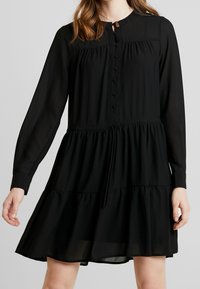 Černé dlouhé šaty s dlouhými rukávy vyrobené z lehkého materiálu. Mají zapínání na knoflíky vpředu, nařasený pas a patrový volánkový lem. Hladká textura.