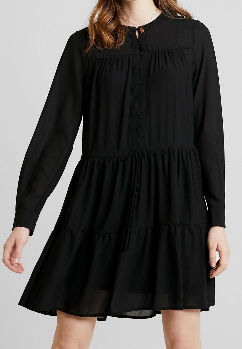 Černé dlouhé šaty s dlouhými rukávy vyrobené z lehkého materiálu. Mají zapínání na knoflíky vpředu, nařasený pas a patrový volánkový lem. Hladká textura.