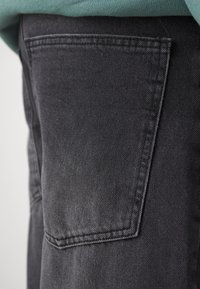 Czarne dżinsy z denimu z tylną kieszenią z widocznym przeszyciem oraz teksturowanym materiałem. Materiał wygląda na lekko zużyty.