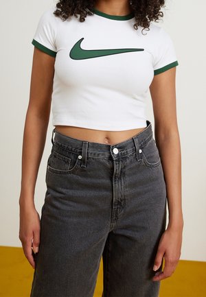 Hvid crop-top t-shirt med grønne detaljer og stort grønt Nike swoosh-logo. Kombinationen inkluderer højttaljede sorte denimjeans.