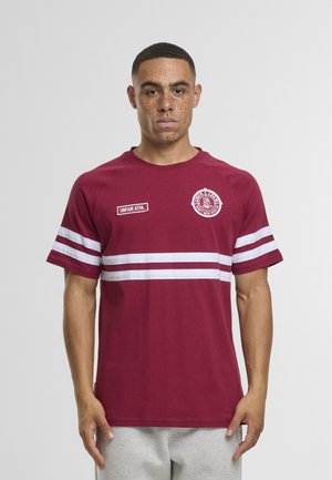 Joven con cabello corto y pecas, vestido con una camiseta color burdeos con rayas blancas y logotipos, posando frente a un fondo gris liso.