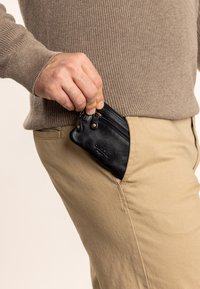 Černá kožená peněženka se zipy, částečně zasunutá do kapsy béžových kalhot. Mezi její vlastnosti patří texturovaná kůže a hardwarové prvky ve zlatém tónu.