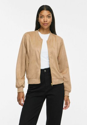 Femme portant une veste beige zippée par-dessus une chemise blanche et un pantalon noir, debout avec une main le long du corps sur un fond blanc.