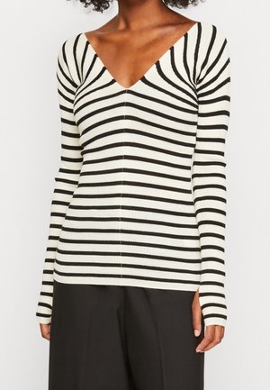 Woman wearing a long-sleeve white sweater with black horizontal stripes and a V-neck, paired with black pants.
