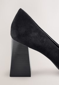 Crna cipelica od brušene kože s blok petom, šiljastim vrhom i prepoznatljivim geometrijskim dizajnom pete. Glatka tekstura i elegantan silueta.