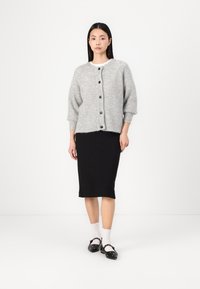 Cardigan gris en tricot avec un col rond et cinq boutons, associé à une jupe crayon noire. Le mannequin porte des chaussettes blanches jusqu'aux chevilles et des chaussures noires.