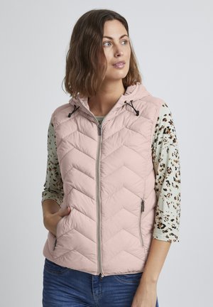 Pink quilted vest with a hood, featuring a chevron pattern, front zipper, and two zipped pockets. Worn over a patterned long-sleeve shirt.