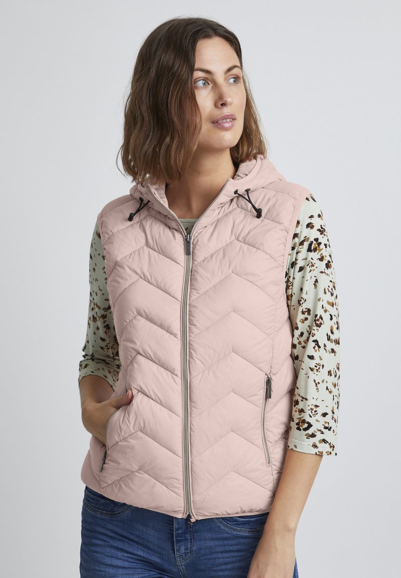 Gilet matelassé rose avec capuche, présentant un motif en chevron, une fermeture éclair à l'avant et deux poches zippées. Porté par-dessus une chemise à manches longues à motif.