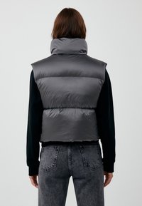 Gilet imbottito grigio con collo alto, caratterizzato da una texture liscia e lucida e un design corto. Indossato sopra una maglia nera a maniche lunghe e jeans.