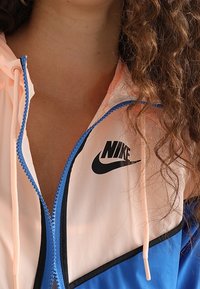 Nike vindjacka med färgblockdesign: pastellrosa, blå och svart. Har dragkedja framtill, huva och signaturlogotyp på bröstet.