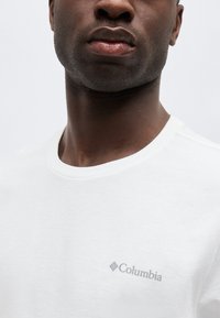 Κοντινό πλάνο ενός άνδρα που φοράει λευκό μπλουζάκι με μικρό γκρι λογότυπο Columbia στο αριστερό μέρος του στήθους, μπροστά σε απλό φωτεινό φόντο.