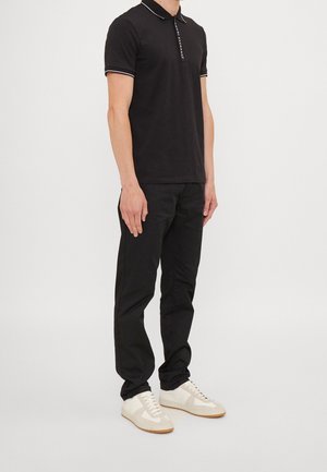 Homme debout portant un polo noir avec bordure blanche, pantalon noir et baskets blanches sur un fond clair uni.