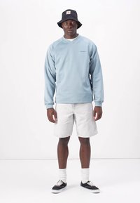 Light blue sweatshirt with long sleeves and a v-neck, paired with light gray shorts. Worn with a black bucket hat and black shoes.