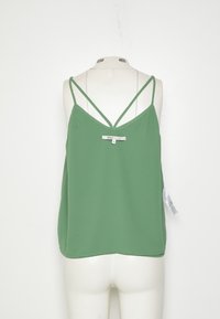 Zelený dámský top bez rukávů s tenkými dvojitými ramínky vystavený na bílém manekýně na jednobarevném pozadí.