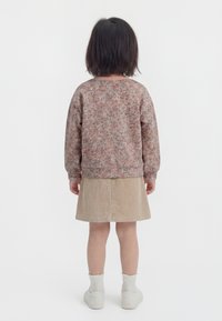 Dziecko z krótkimi ciemnymi włosami, ubrane w różowy sweter w kwiaty, beżową sztruksową spódnicę, białe skarpetki i białe buty, stojące tyłem.