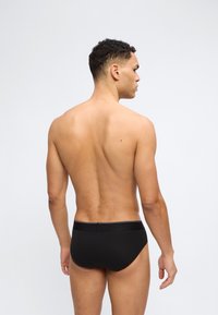 Άνδρας με κοντά σγουρά μαλλιά, γυρισμένος προς τα πίσω, φοράει μαύρα εσώρουχα, με φόντο ένα απλό ανοιχτό γκρι φόντο.