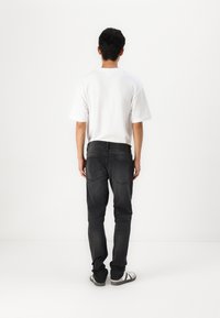 Hvit kortermet t-skjorte, svarte jeans med en teksturert overflate, og svart-hvite joggesko, sett fra baksiden på en ensfarget bakgrunn.
