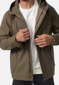 Chaqueta verde oliva con capucha, detalles de cordón y cremallera frontal; lleva puesta sobre un sudadera blanca con acentos texturizados y pantalones negros.