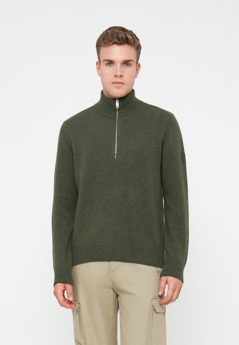 Mladý muž v olivovo zelenom zipsovom svetri a béžových cargo nohaviciach, stojaci pred čistým bielym pozadím.