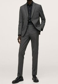 Grau maßgeschneiderter Anzug mit passformbetonter Gestaltung und subtiler Nadelstreifenstruktur, kombiniert mit einem schwarzen Knopfhemd und schwarzen Schuhen.