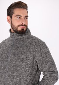 Giacca in pile grigia con zip e colletto alto. Presenta un motivo melange testurizzato e dettagli di cucitura sottili lungo la cerniera.