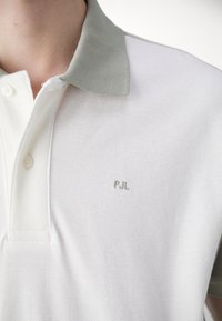 Biela polokošeľa so svetlozeleným golierom, textúrovaná látka a vyšité logo "PJL" v zodpovedajúcej zelenej farbe na ľavej strane hrudníka.