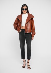 Chaqueta de cuero marrón corta con cuello ancho, top corto blanco, jeans negros de talle alto con botón frontal y tacones negros con tiras.