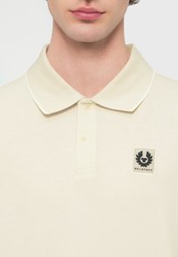 Μπεζ πόλο πουκάμισο με υφαντό υφή, διαθέτει κλασικό γιακά, πόρπη με δύο κουμπιά και μαύρο λογότυπο Belstaff στο στήθος.