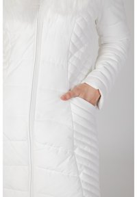 Manteau en duvet blanc avec un design matelassé, fermeture éclair sur le devant et col en fausse fourrure. Dispose de poches latérales et d'une texture lisse et brillante.