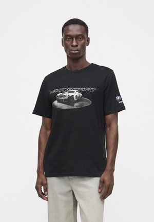 Mand iført sort t-shirt med "MOTORSPORT" tekst og en bilgrafik på fronten samt et BMW-logo på højre ærme, kombineret med lysegrå bukser.