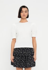 Haut blanc texturé à manches courtes bouffantes, porté avec une jupe noire à volants ornée d'un motif floral. Encolure ronde simple.