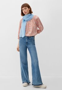 Bluse mit floralem Muster in Orange und gerafftem Design, kombiniert mit hoch taillierten, weit geschnittenen blauen Jeans. Ein hellblauer, strukturiertes Schal legt sich um den Hals.