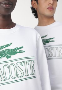 Due persone che indossano felpe bianche con il logo verde Lacoste che presenta un coccodrillo e il nome del marchio.