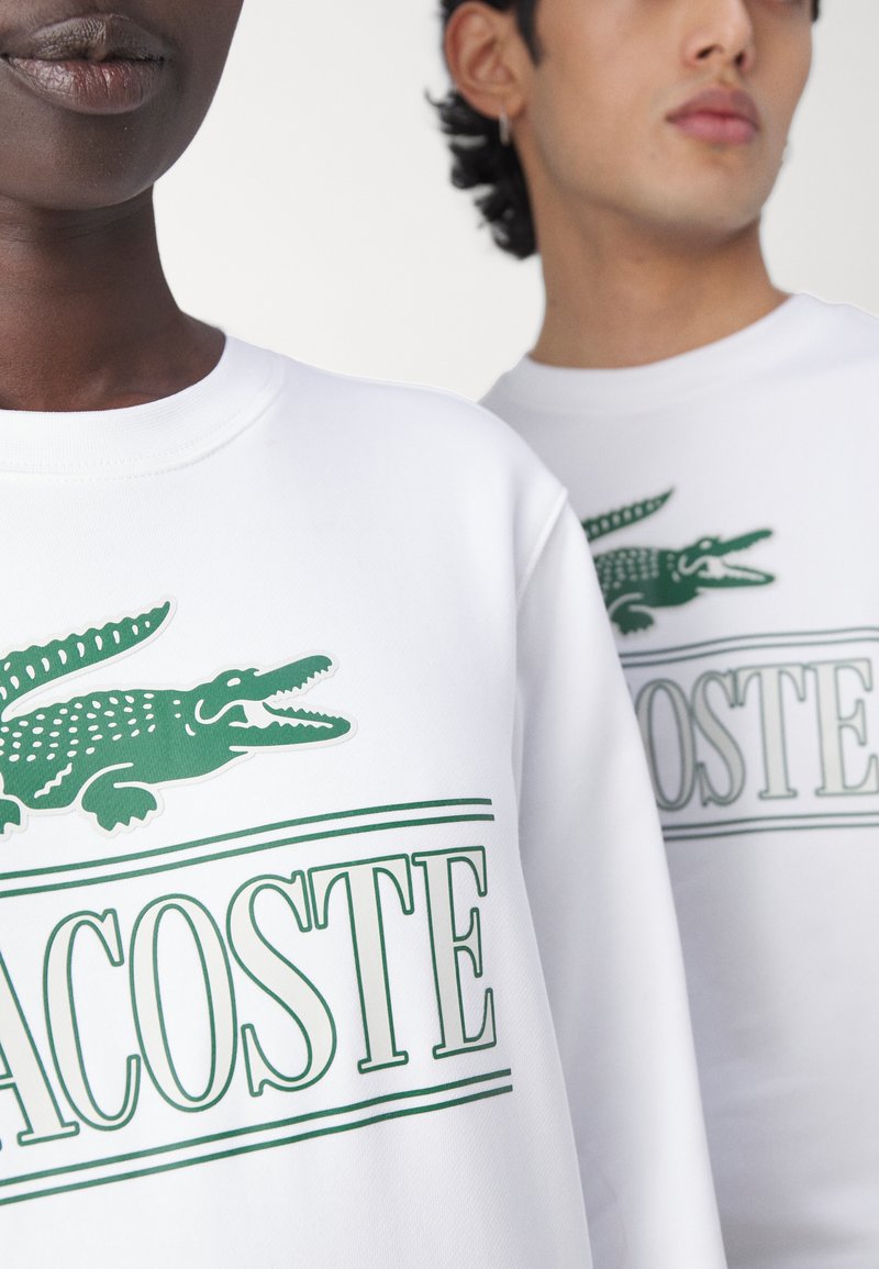 Dos personas llevan sudaderas blancas con el logo verde de Lacoste, que presenta un cocodrilo y el nombre de la marca.