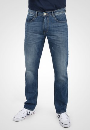 Jeans in denim blu con taglio dritto, dotati di cinque tasche, chiusura con bottone e sbiaditura visibile sul davanti. Indossati con sneakers bianche.