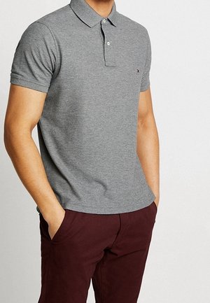 Homme portant un polo gris à manches courtes et un pantalon rouge foncé, les mains dans les poches, sur un fond blanc uni.