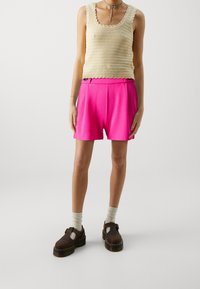 Débardeur beige en maille avec bords festonnés, associé à un short rose vif. Chaussures marron à semelle épaisse avec détails à bride et chaussettes crème.