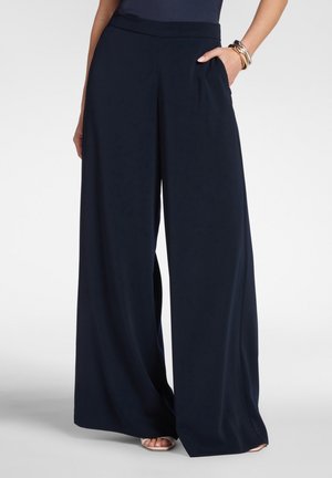 Donna che indossa pantaloni neri a gamba larga con tasche, abbinati a tacchi open-toe e braccialetti al polso sinistro, in piedi contro uno sfondo bianco.