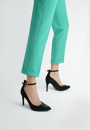 Scarpe nere con tacco alto, punta affusolata, cinturino alla caviglia e dettagli in strass decorativi, abbinate a pantaloni teal a risvolto.