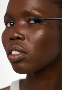 Zbliżenie na twarz nakładającą niebieską maskarę, podkreślającą gładką ciemną skórę, pełne usta i naturalny, matowy wygląd. Widoczne subtelne rysy twarzy.