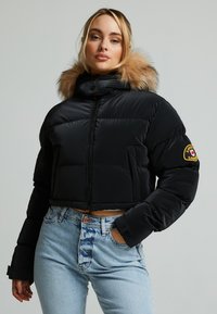 Zwarte cropped pufferjack met een capuchon met bonttrim, ritsluiting en logo-opgestikte patch op de mouw. Gecombineerd met high-waisted vervaagde blauwe jeans.