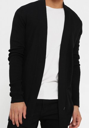 Hombre con un cárdigan negro abierto sobre una camiseta blanca y pantalones negros, de pie con las manos relajadas a los lados.
