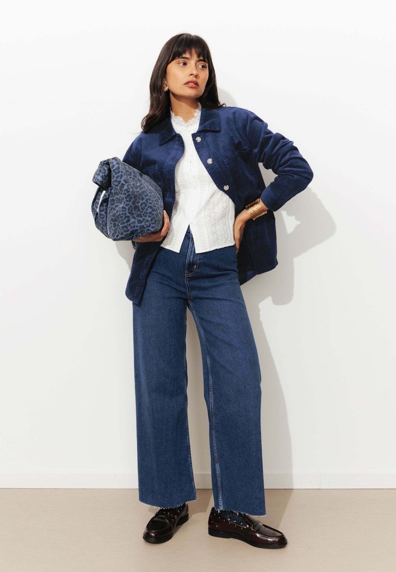 Veste en velours côtelé bleu sur un blouse en dentelle blanche, associée à un jean en denim taille haute et des mocassins noirs. Tient un sac à motifs léopard bleu.