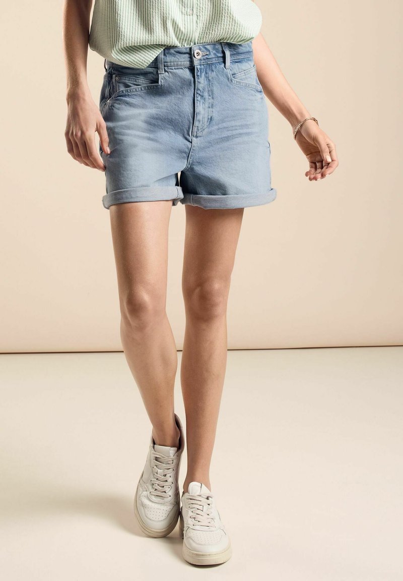 Pantalones cortos de denim azul claro con dobladillos enrollados, que cuentan con bolsillos frontales y cierre de botón, combinados con zapatillas blancas sobre un fondo neutro.
