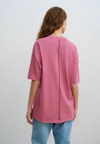 Lyserød bomulds-t-shirt med korte ærmer og en afslappet pasform. Har en centered rynke på ryggen og en rund halsudskæring. Sammen med blå jeans.