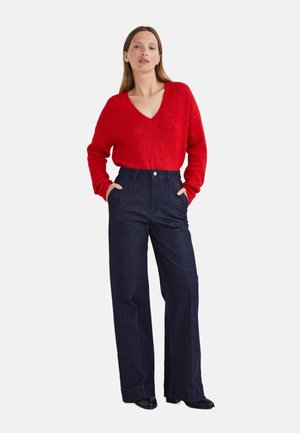 Pull rouge en V fabriqué dans un matériau doux, associé à un pantalon large taille haute en denim foncé avec des poches avant et une fermeture à bouton.