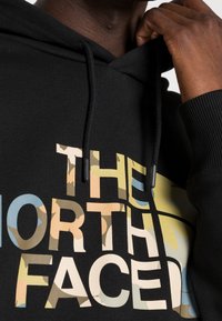 Felpa nera con cappuccio e una grafica frontale che presenta "THE NORTH FACE" in beige, blu e motivi camouflage. Include un cappuccio con cordino e polsini a coste.