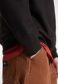 Sweatshirt noir en tissu texturé avec logo en relief sur le poignet, superposé à une chemise rouge et un pantalon marron avec une étiquette.